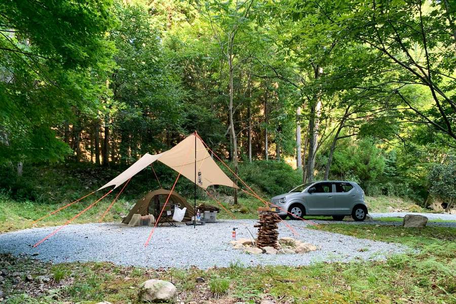 森の中にある区画の中にタープとテントが貼られている。横には車が停まっている。