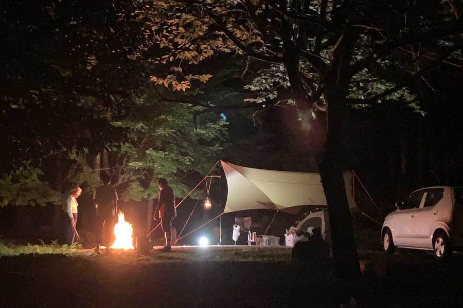 夜、森の中にある区画の中で焚き火をしている画像。焚き火の横に２人の大人が立っている。横にタープとテント、車が写っている。
