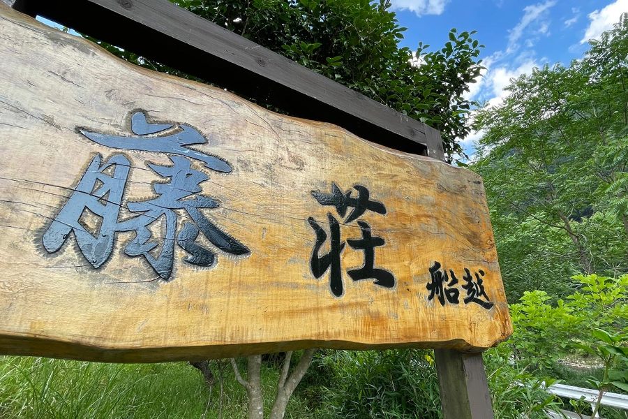 藤荘キャンプ場の入り口にある看板を寄りで撮った画像。木材の板に「藤荘 船越」と書かれている。後ろに森と青空が見える。