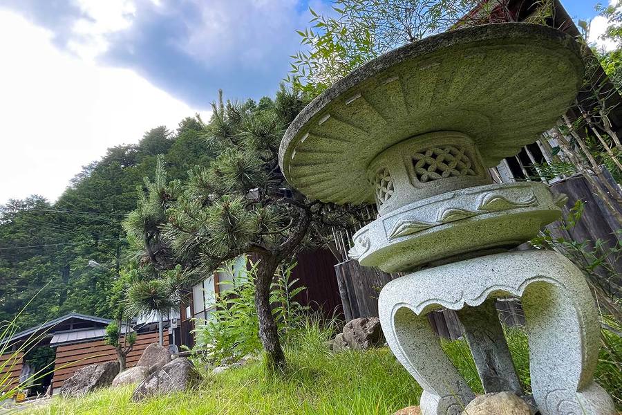 晴れた空の下、森の中にある共同水場の前に重厚感のある石の飾りが見える。