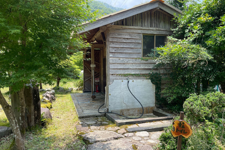 共同トイレを外から撮影した画像。晴れた空の下、森の中に小屋（共同トイレ）が見える。小屋の外側にはホースが設置されている。