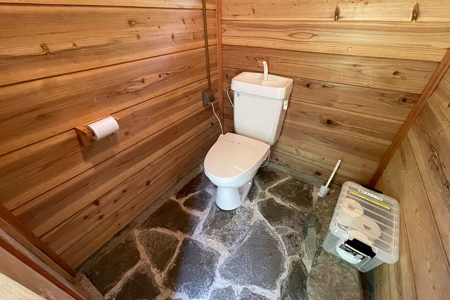 トイレの画像。小屋の扉を開けると水洗トイレとトイレットペーパーが見える。石でできた床材、掃除用具と、容器に入ったトイレットペーパーが見える。。