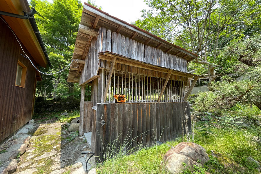 共同水場を外から撮影した画像。晴れた空の下、森の中に小屋（共同水場）が見える。